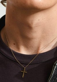 Collana con croce d'oro su una sottile catenina, appoggiata su una superficie cutanea testurizzata, sopra un tessuto a coste scuro del colletto.