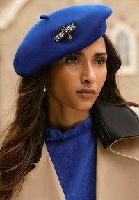 Zils vilnas berets ar dekoratīvu melnu un caurspīdīgu rhinestone piespraudi. Dizains ietver noapaļotu formu un mīkstu tekstūru.