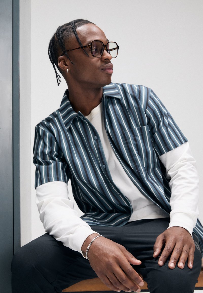 Πουκάμισο με κοντά μανίκια και κουμπιά, με κάθετες ρίγες σε μπλε και άσπρο, συνδυασμένο με λευκό πουλόβερ με μακριά μανίκια και σκούρο παντελόνι.