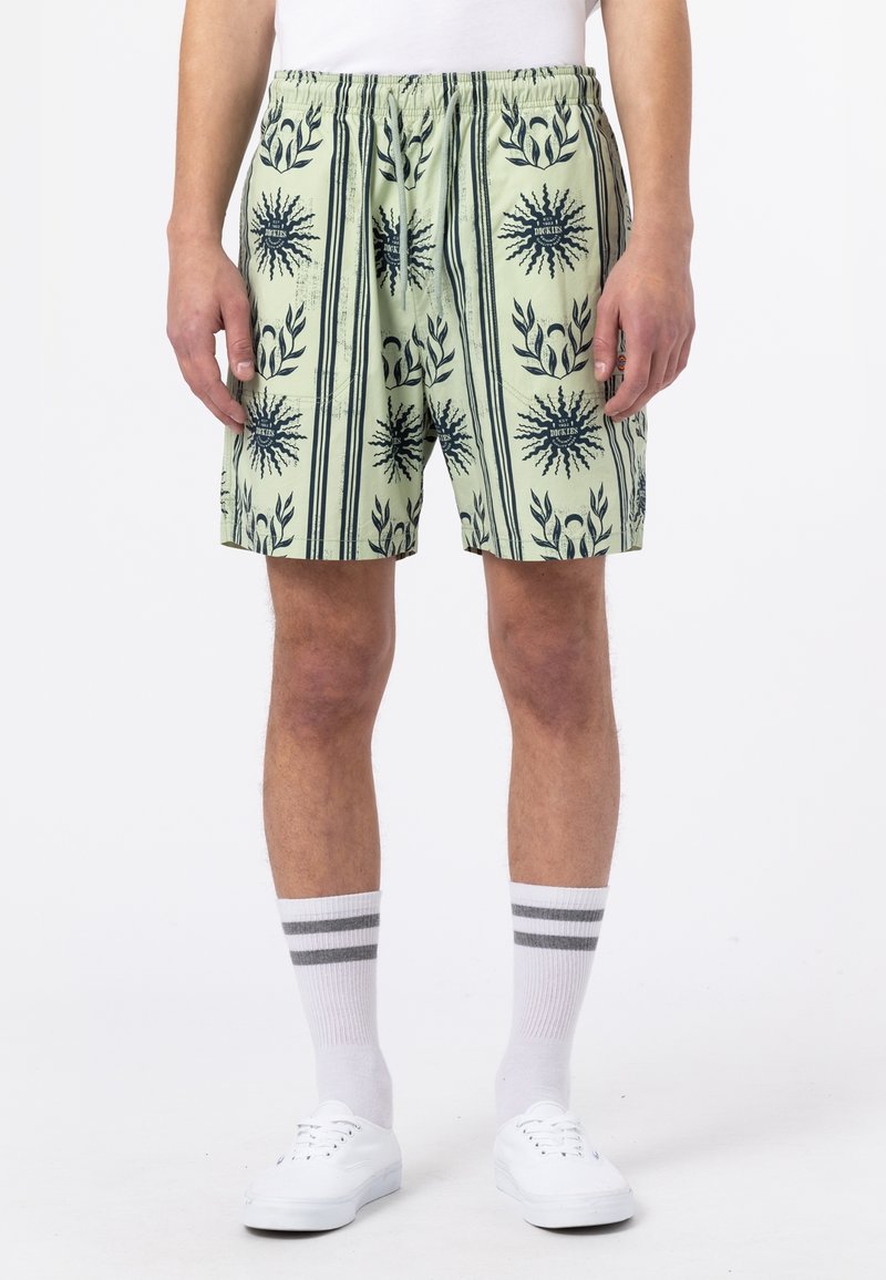 Grüne gemusterte Shorts mit schwarzen Blatt- und Sonnendesigns, elastischem Bund und Seitentaschen; kombiniert mit weißen Socken und Sneakers.