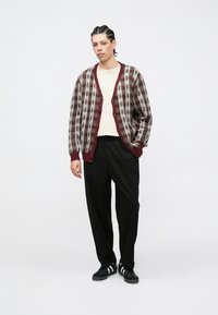 Cardigan en laine tricotée à carreaux bordeaux et gris, boutonné devant, coupe décontractée. Associé à un pantalon noir et des baskets, avec une chemise crème en dessous.