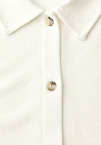 Camiseta blanca de punto con cuello puntiagudo y dos botones de mármol beige abrochados en la tapeta frontal.