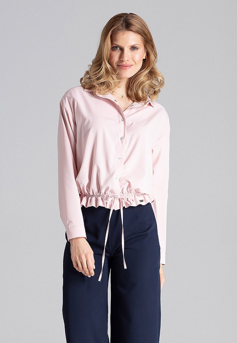 Jasnoróżowa, długofalowa, krótka koszula z długim rękawem z zapięciem na guziki z przodu i ściągaczem na dole. Wykonana z gładkiego materiału o luźnym kroju.