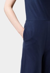 Granatowy kombinezon wykonany z gładkiej tkaniny, z szerokimi nogawkami oraz bocznymi kieszeniami. Górna część płynnie przechodzi w dolną.