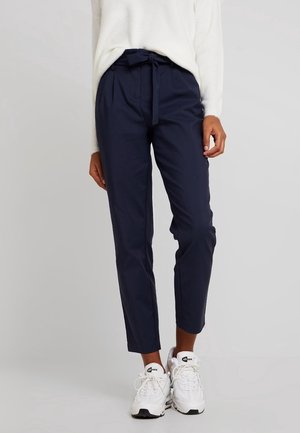 Persona con pantalones paperbag de talle alto en azul marino con cinturón atado, suéter blanco y zapatillas Air Max blancas, de pie sobre un fondo gris liso.