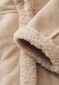 Tissu doux beige avec une doublure en polaire crème, moelleuse et texturée le long du col et des poignets. Matériau extérieur lisse avec un subtil éclat.