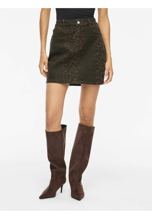 Femme portant une mini-jupe imprimé animalier marron foncé et des bottes à talons en suède marron montant jusqu’aux genoux, debout, mains relâchées sur les côtés.