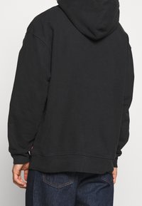 Sweat à capuche noir avec une coupe décontractée, une texture douce et des poignets côtelés. Dispose d'une poche kangourou et d'une discrète marque près de l'ourlet.