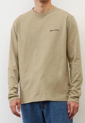 Mężczyzna w beżowej, długorękawowej koszuli Marc O’Polo i niebieskich dżinsach, stojący na tle gładkiej, białej ściany.