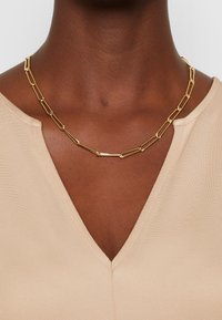Collier chaîne en or avec des maillons rectangulaires allongés porté sur un cou à peau foncée, sur un haut beige à col en V.