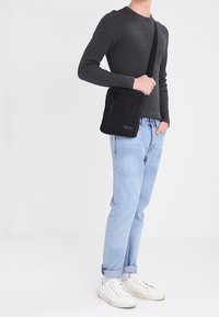 Persona che indossa un maglione grigio, jeans azzurri chiari, scarpe da ginnastica bianche e porta una borsa a tracolla nera con una tasca frontale con zip.