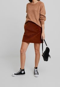 Pull en tricot marron, jupe en velours côtelé rouille, baskets montantes noires et sac à bandoulière noir avec une finition texturée.