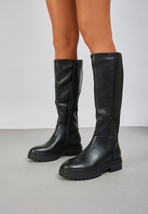 Bottes noires en cuir montantes jusqu'aux genoux, avec une texture lisse, dotées d'une fermeture éclair sur le côté, d'une semelle en caoutchouc épaisse et de détails en tissu côtelé.