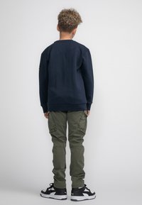 Felpa navy con colletto a girocollo, pantaloni cargo verdi con tasche laterali e sneakers bianco e nere. Bambino visto da dietro, capelli ricci visibili.