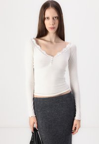 Langermet hvit ribbet topp med bølget blondekrage og knappedetaljer. Kombinert med en mørkegrå tettsittende ribbet skjørt.