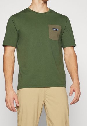 Homme portant un t-shirt vert olive à manches courtes avec une poche poitrine contrastante et un pantalon beige sur fond uni.