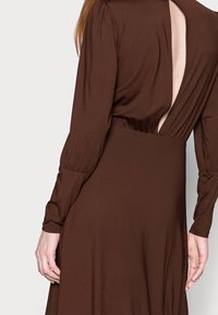 Brun, långärmad klänning med veckad detaljer på ryggen och mjuk textur. Tyget faller smidigt, med en figurnära midja och en svepande kjol.