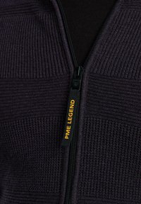 Veste zippée violet foncé avec panneaux texturés, dotée d'une fermeture éclair noire et d'une étiquette "PME LEGEND" jaune sur le ruban de la fermeture.