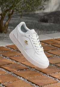 Lacoste balta sportinė avalynė su lygiu odiniu paviršiumi, tekstūruotu guminiu padu ir auksiniu logotipu. Turi apvalų priekį ir klasiką raištelių dizainą.