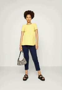 Camisola amarela de manga curta com bainha assimétrica, combinada com jeans azul escuro e sandálias plataforma pretas. Segurando uma mala de mão com padrão de zebra em preto e branco.