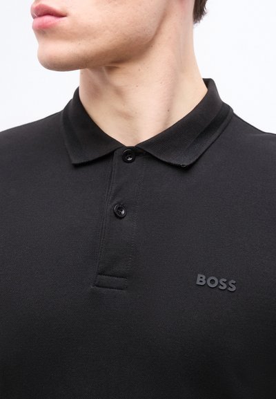 Gros plan d'une personne portant un polo noir à deux boutons avec le logo "BOSS" sur la poitrine, regardant légèrement sur le côté.