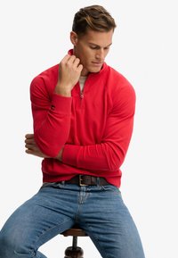 Pull en polaire rouge à zip quart avec un col haut et des poignets côtelés, associé à un jean bleu et une ceinture brune, assis sur un tabouret.