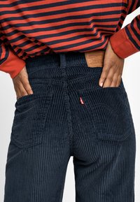 Personne portant un pantalon en velours côtelé bleu marine avec les mains dans les poches arrière, associée à un t-shirt à manches longues rayé rouge et bleu marine.