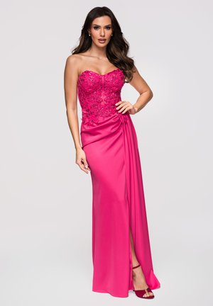 Frau mit langen dunklen Haaren, die ein trägerloses fuchsiafarbenes Abendkleid mit Blumenstickerei und seitlichem Schlitz trägt, posiert mit einer Hand auf der Hüfte.