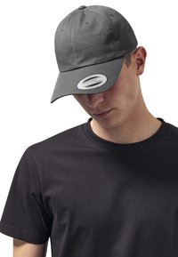 Szürke pamut baseball sapka ívelt szélekkel, elöl egy kör alakú címkével. Fekete kerek nyakú pólóval viselve.