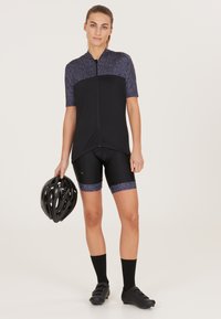 Svart cykeltröja med korta ärmar, med ett mörkt mönstrat övre avsnitt. Svarta vadderade shorts med mönstrade detaljer. Svart hjälm i handen.