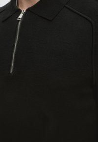 Homme portant un polo en maille noire avec un col court et une demi-fermeture éclair argentée sur la poitrine, visible du cou à la moitié du torse.