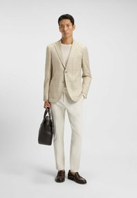 Beige geruite blazer met twee voorkruiden, gecombineerd met crèmekleurige broeken. Bruine leren schoenen en een donkere tas maken de outfit compleet.