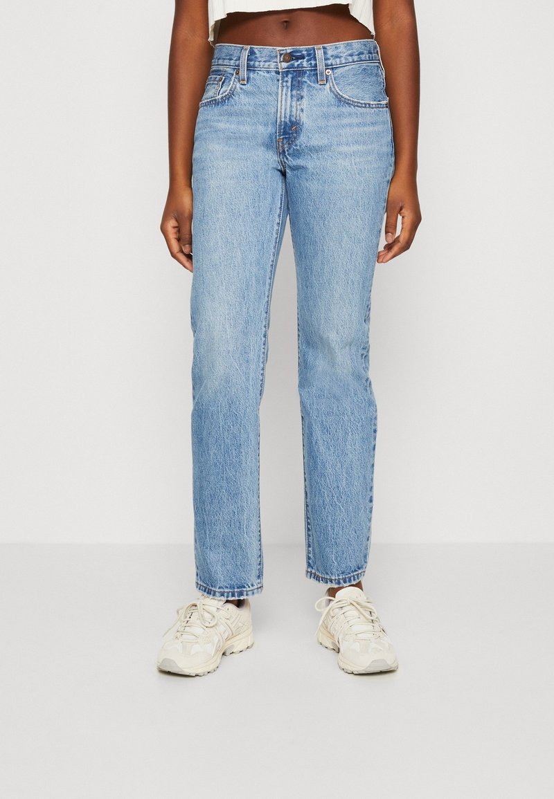 Femme portant un jean bleu clair droit et des baskets blanches, debout devant un fond blanc uni.