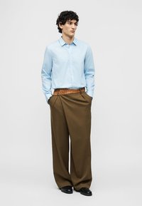 Homme aux cheveux bouclés portant une chemise bleu clair, un pantalon large marron avec ceinture, des chaussures noires, debout, les mains dans les poches, regardant sur le côté.