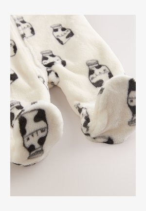 Une combinaison en polaire couleur crème avec un imprimé vache noir et des motifs de cruche à lait, offrant une texture douce et des pieds fermés pour plus de chaleur.