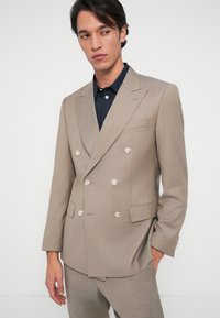 Μπεζ σακάκι κοστούμι διπλής όψης με δομημένο σχέδιο, με αυχένες σε προεξοχή και κρέμ κουμπιά, σε συνδυασμό με σκούρο μπλε πουκάμισο από κάτω.