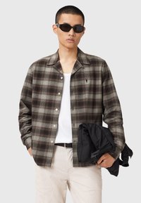 Man wearing sunglasses, brown plaid button-up shirt over white tank top, beige pants, holding folded black jacket against gray background.