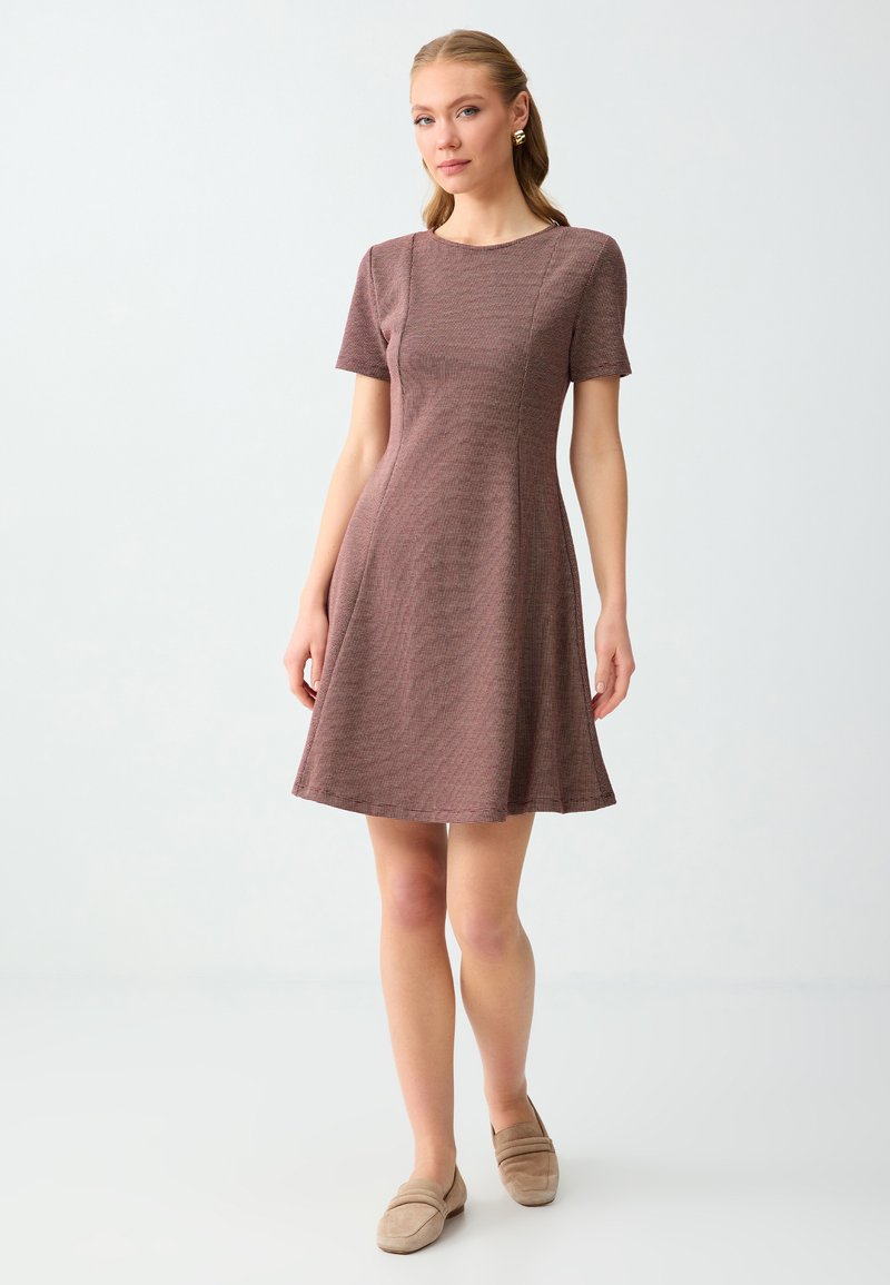 Femme debout portant une robe en maille mauve à manches courtes et des mocassins beiges sur un fond gris clair uni.