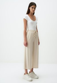 Beige wijde broek met een elastische tailleband en trekkoorddetails, gecombineerd met een aansluitend wit geribd topje en witte sneakers.