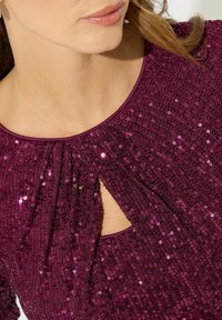 Top viola vivace con paillettes, caratterizzato da scollatura a cerchio e un dettaglio a chiave centrale, che mette in risalto il tessuto testurizzato e l'eleganza delle pieghe.