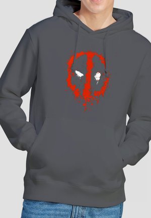 Person trägt einen dunkelgrauen Hoodie mit einem rot-weißen stilisierten Deadpool-Logo auf der Brust und blaue Jeans. Hände in den Taschen.