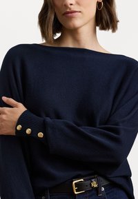 Tengerészkék kötött pulóver széles, hajónyakú körvonalakkal és bordázott mandzsettákkal, három aranygombos részlettel. Fekete övvel párosítva.