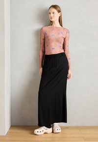 Longue jupe noire avec une fente sur le côté, associée à un haut ajusté à manches longues de couleur rose clair avec des motifs abstraits bleus ; sabots blancs.
