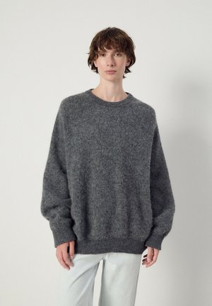Persona con capelli corti castani che indossa un maglione oversize grigio scuro e morbido e pantaloni chiari, in piedi davanti a uno sfondo chiaro uniforme.