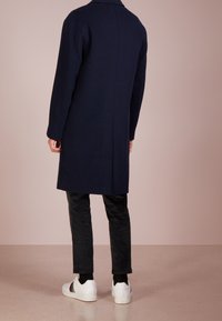 Dunkelblauer Wollmantel mit geradem Schnitt, Rückennahtdetails und langen Ärmeln, kombiniert mit schwarzen Hosen und weißen Sneakers.