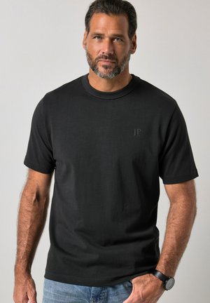 Uomo che indossa una maglietta nera a maniche corte e jeans blu, con un orologio da polso al polso sinistro, in piedi davanti a uno sfondo chiaro e uniforme.