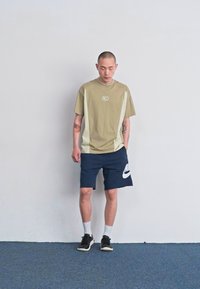 Tričko s krátkym rukávom v olivovo zelenej farbe s bielymi bočnými panelmi, námornícke šortky so bielym logom Nike a čierne športové topánky.