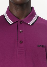 Lila Poloshirt aus strukturiertem Material, mit einem gestreiften Kragen in Weiß und Schwarz, drei Knöpfen und einem gestickten "BOSS"-Logo.