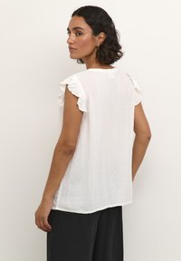 Haut blanc léger avec des manches volantes, motif texturé et finition douce. Le dos présente une petite fermeture au cou et une coupe décontractée.