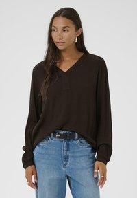 Camicia lunga marrone e strutturata con scollo a V e vestibilità ampia, abbinata a jeans blu a vita alta e una cintura nera.